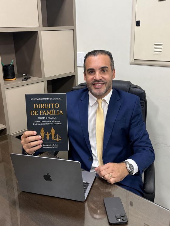 Rosevaldo Suzart de Oliveira sentado à mesa do escritório, de terno azul, segurando o livro Direito de Família: Teoria e Prática, que ele escreveu.