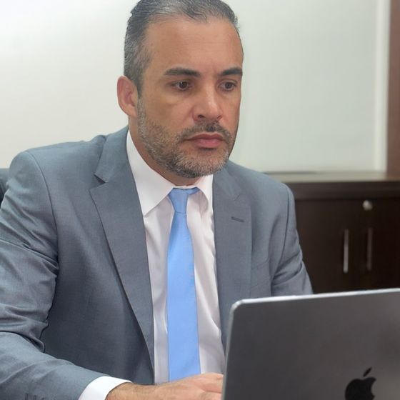 Rosevaldo Suzart de Oliveira de terno cinza, sentado à mesa do escritório, trabalhando no computador durante uma audiência online.